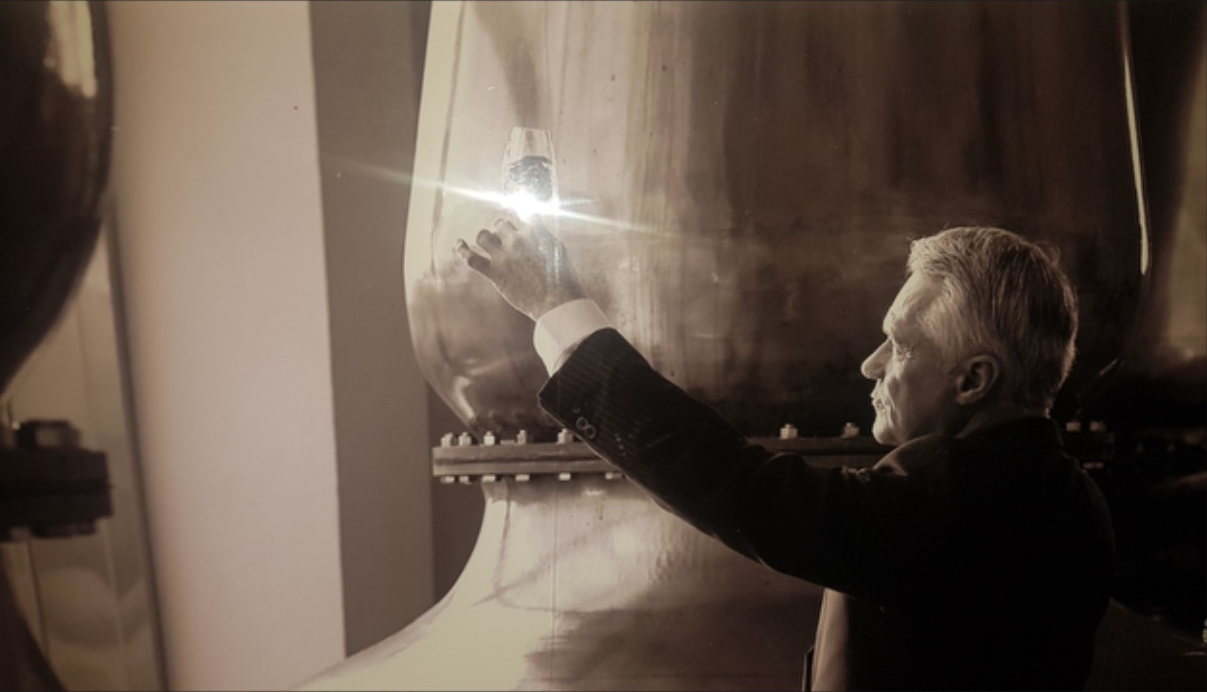 Dr Jim Swan holding a glass of spirit up to the light beside a still at a malt distillery.